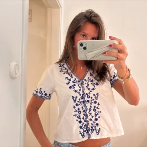 Vit blus med blå broderier från Zara - Superfin vit blus från Zara med blå broderade blommor och v-ringning. Korta ärmar med broderade detaljer och avslappnad passform. Perfekt till jeans eller kjol.