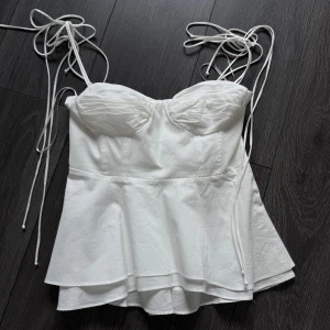Vit korsettopp med knyt från Zara - Supersöt vit korsettopp från Zara med tunna axelband som knyts och markerade kupor. Toppen har en lätt utsvängd peplum-nederdel och är tillverkad i ett mjukt tyg som känns lyxigt. Perfekt för dig som gillar romantiska och trendiga detaljer. Den är helt oanvänd utan lappen (har dock provat på den. Men den passade tyvärr inte) Ord pris = 400
