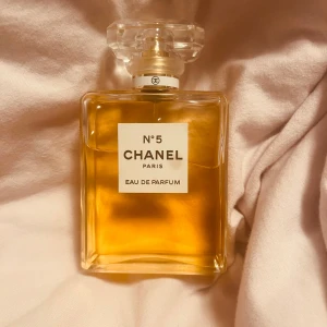 Chanel No 5 Eau de Parfum 100ml - Klassisk Chanel No 5 Eau de Parfum i 100 ml flaska. Parfymen kommer i en ikonisk, rektangulär glasflaska med transparent, multifacetterad kork och guldig vätska. Elegant och tidlös design med tydlig Chanel-logga på etiketten och korken.