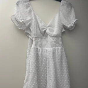 Vit byxdress med puffärm - Superfin vit byxdress med puffiga kortärmar och v-ringning. Klänningen har ett sött prickigt strukturmönster och smock i midjan för en bekväm passform. Öppen i ryggen men knyte för en elegant och söt look. Perfekt för sommarens alla tillfällen!