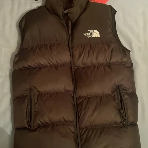 Svart dunväst från The North Face S - Säljer en svart dunväst från The North Face i storlek S. Västen är quiltad med hög krage, dragkedja framtill och två fickor med dragkedja. Klassisk logga broderad på bröstet och ryggen. Perfekt för lager-på-lager under kyliga dagar.