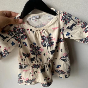 Beige klänning med djurmönster Polarn o. Pyret barn 74 - Supersöt beige kortklänning från Polarn o. Pyret med blått och rosa djurmönster, träd och svampar. Klänningen har lång ärm, rund hals och praktiska fickor framtill. Tillverkad i mjuk bomull, perfekt för lekfulla dagar.