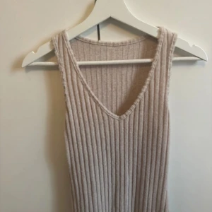 Beige ribbad långklänning - Superfin beige långklänning i ribbat material med v-ringning och ärmlös design. Klänningen är stickad och har en mjuk, skön känsla som passar perfekt för en avslappnad men ändå stilren look.