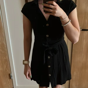 Svart kort klänning från H&M - Svart kort klänning från H&M med knappar framtill och knytskärp i midjan. Klänningen har krage, v-ringning. Viskos och linne blandning 