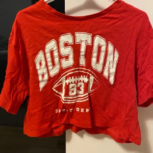 Röd croppad Boston-topp H&M - Cool röd croppad topp från H&M med stort vitt Boston-tryck och amerikansk fotbollsgrafik framtill. T-shirten har korta ärmar och avslappnad passform, perfekt för en sportig look. Tillverkad i mjuk bomull. Skrynklig på bilden men fint skick! 