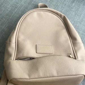 Beige ryggsäck i skinnimitation - Snygg beige ryggsäck från Don Donna i skinnimitation med justerbara axelband och guldfärgade dragkedjor. Väskan har en stilren design med ett huvudfack och en mindre ficka framtill, perfekt för vardagens alla prylar.