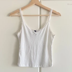 Vit spetskantad linnetopp - Supersöt vit linnetopp med smala axelband och spetskant längs ringningen och banden. Toppen har en liten rosett framtill och ett diskret mönster i tyget som ger en romantisk känsla. Perfekt att styla med jeans eller kjol för en fräsch look.