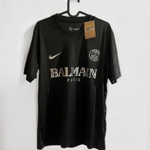 Svart PSG x Balmain fotbollströja Nike - Snygg svart fotbollströja från Nike i samarbete med PSG och Balmain. Tröjan har korta ärmar, rund hals och tryck i silver på bröstet med Balmain Paris och PSG-logga. Materialet är lätt och andas, perfekt för träning eller match.