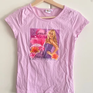 Rosa Hannah Montana t-shirt - Söt rosa t-shirt med tryck av Hannah Montana och texten 'style to spare' framtill. Klassisk rund halsringning och korta ärmar. Perfekt för dig som älskar nostalgi och popkultur. Mjuk och skön i bomull.