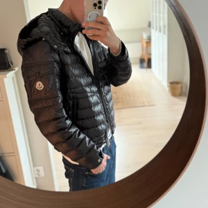 Svart Moncler dunjacka med huva - Säljer en svart Moncler dunjacka med huva och dragkedja. Jackan har klassisk quiltad design, Moncler-logga på ärmen och är riktigt lätt och smidig. Perfekt för dig som vill ha en stilren och varm jacka med premiumkänsla.