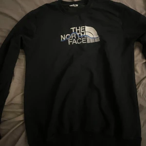 Svart sweatshirt från The North Face - Svart sweatshirt från The North Face med stort broderat logotyptryck i silver och blått på bröstet. Klassisk rund halsringning och långa ärmar. Perfekt för en chill och stilren look.
