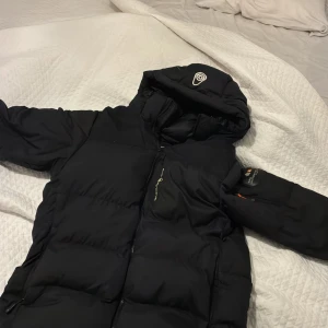 Svart Sail Racing dunjacka M - Säljer en svart dunjacka från Sail Racing i storlek medium. Jackan har en rejäl huva med broderad logga, dragkedja framtill och flera praktiska fickor. Perfekt för kalla dagar och riktigt snygg streetstil.