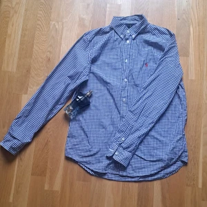 Rutig blå skjorta från Ralph Lauren - Klassisk rutig skjorta från Ralph Lauren i blått och vitt med röd broderad logga på bröstet. Skjortan har lång ärm, knappar hela vägen och button-down krage. Tillverkad i mjuk bomull och passar perfekt till jeans eller chinos. Pris 299 kr