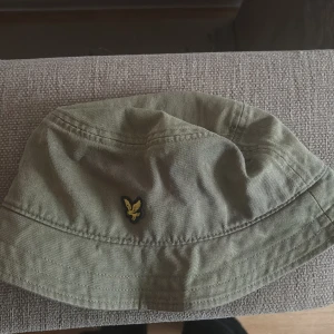 Olivgrön bucket hat från Lyle & Scott - Bucket hat i olivgrön färg från Lyle & Scott med klassisk gul broderad fågel framtill. 