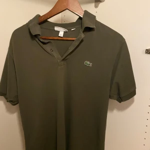 Olivgrön Lacoste pikétröja - Snygg olivgrön piké från Lacoste med klassisk krage och två knappar framtill. Ikonisk broderad krokodil på bröstet. T-shirten är gjord i bomull och har korta ärmar, perfekt för chill dagar. Passar dig som gillar stilren och sportig vibe.