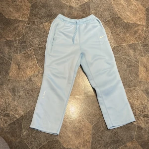 Nike NOCTA ljusblå mjukisbyxor M - Snygga ljusblå mjukisbyxor från Nike NOCTA med dragsko i midjan och diskret NOCTA- och Swoosh-logga. Byxorna har raka ben, dragkedjeficka och är gjorda i mjukt bomullsmaterial. Perfekta för chill eller streetwear.
