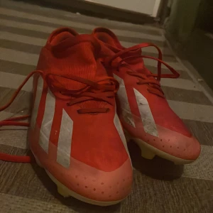 Adidas Crazyfast röda fotbollsskor - Säljer ett par Adidas Crazyfast fotbollsskor i rött med vita detaljer och klassiska tre ränder på sidorna. Skorna har snörning och platt sula med dobbar för bra grepp på planen. Lätta och smidiga för snabba rörelser. Perfekta för dig som vill sticka ut på fotbollsplanen.