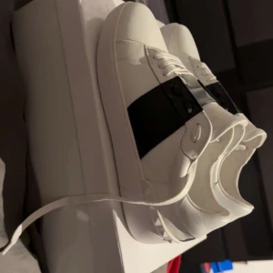 Valentino skor  - Snygga Valentino Open sneakers i vitt skinn med bred svart läderdetalj över mitten. Klassisk rund tå, vita snören och ikoniska nitar baktill på sulan. Perfekta för dig som gillar stilrena och lyxiga sneakers med en twist.