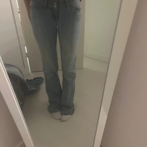 Ljusblå bootcut jeans med broderi - Säljer ett par ljusblå jeans med bootcut-modell och snyggt broderi i rosa på bakfickorna. Jeansen har normal passform och är tillverkade i mjukt denimtyg. Midjemåttet är 30 cm och innebrenslängden är 84 cm. Dom har lite missfärgningar längst ner på byxorna