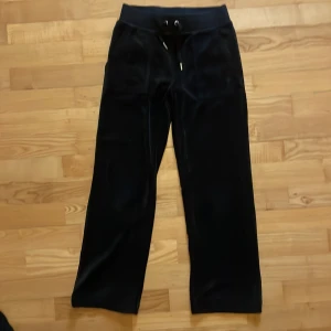 Svarta velourbyxor Juicy Couture - Mysiga svarta mjukisbyxor från Juicy Couture i glansig velour. Byxorna har bred resår i midjan med snörning, raka ben och fickor både fram och bak med snygga knappar. Klassisk JC-logga broderad på ena fickan framtill. Benlängd 85 cm.