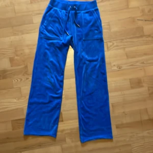 Blåa Juicy Couture mjukisbyxor - Snygga blåa mjukisbyxor från Juicy Couture med bred resår i midjan och snörning. Byxorna har raka ben, stora fickor fram och bak med silvriga knappar samt broderad JC-logga på ena fickan. Mjukt material som känns lyxigt. Byxorna är uppsydda och benlängden är 85 cm.