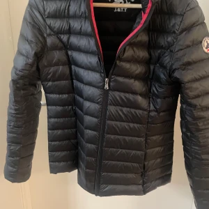 Svart dunjacka från JOTT med dragkedja - Säljer en svart dunjacka från JOTT med horisontella sömmar och röd kant längs insidan. Har använt den förra hösten och vill sälja för den är för stor. Köpte den i butik och jackan kommer även med en påse som man kan lägga den i.