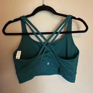 Grön sport-bh från Lululemon - Supersnygg grön sport-bh från Lululemon med korsade axelband i ryggen. Perfekt för träning eller yoga, tillverkad i mjukt och stretchigt material som andas. Enkel och stilren design med diskret logga framtill. Sitter skönt och ger bra stöd.