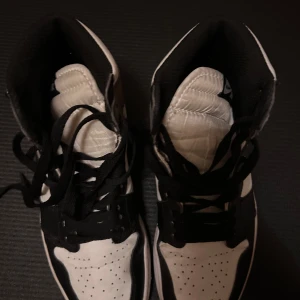 Nike Air Jordan 1 High svart/vit - Säljer ett par Nike Air Jordan 1 High i svart, vitt och grått. Klassisk high-top siluett med svart swoosh, svarta skosnören och Air Jordan-logga på sidan. Ovandel i skinn och syntet, perforerad tå och vit mellansula. Perfekta för dig som gillar streetstyle.