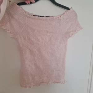 Ljusrosa smockad offshoulder topp - Supersöt ljusrosa topp med smockad struktur och offshoulder-modell. Korta ärmar och volangkant vid hals, ärmslut och nederkant. Perfekt för en somrig look och enkel att matcha med jeans eller kjol.