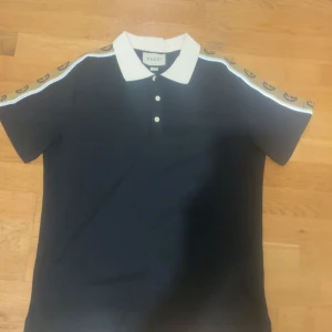 Svart Gucci t-shirt med logoband - Snygg svart t-shirt från Gucci med vit krage och knappar framtill. Ärmarna har beige band med Gucci-loggor längs axlarna för en extra lyxig vibe. Perfekt för dig som vill sticka ut med stilren design och exklusiva detaljer.