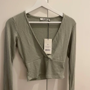 Ribbad omlottopp från NA-KD grön XS - Snygg ribbad omlottopp från NA-KD i ljusgrön nyans. Toppen har lång ärm, croppad passform och djup v-ringning. Perfekt att styla med höga jeans eller kjol för en trendig look.