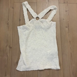 Vit smockad linnetopp med ringdetalj - Supersöt vit linnetopp med smockat tyg och breda axelband som möts i en ringdetalj bak. Toppens struktur ger en cool och avslappnad vibe, perfekt till jeans eller kjol. Lätt och luftig för varma dagar.