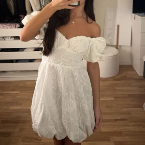 Vit kortklänning från Viva Couture-New Yorker  - Supersöt vit kortklänning från Viva Couture med puffiga offshoulder-ärmar och broderade blommor. Klänningen har en voluminös kjol och markerad midja, perfekt för studenten samt midsommar. Materialet känns lätt och luftigt som är bra för en varm sommardag. Har använts en gång på min student mottagning, alltså har den inte fått någon champagne på sig eller liknande. 