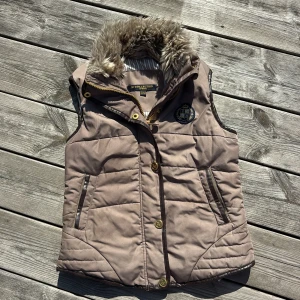 Beige dunväst med fuskpäls från JH Collection 134/140 - Snygg beige dunväst från JH Collection med mjuk fuskpäls i kragen och guldfärgade detaljer. Västen har broderat emblem på bröstet, dragkedja och knappar framtill samt två fickor. Insidan av kragen har randigt tyg och baktill finns dekorativa dragkedjor.