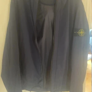 Blå vindjacka Stone Island M - Snygg marinblå vindjacka från Stone Island med klassisk patch på ärmen. Jackan har huva, dragkedja framtill och är tillverkad i lätt polyester. Perfekt för dig som vill ha en clean och sportig look med streetkänsla.