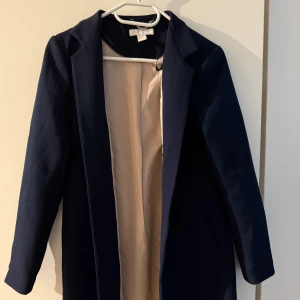Mörkblå kappa från HM - Stilren mörkblå kappa från Hm med klassisk krage och helfodrad insida i beige. Rak passform och långa ärmar, perfekt för dig som vill ha en clean och tidlös look. Passar till allt och ger en snygg siluett.