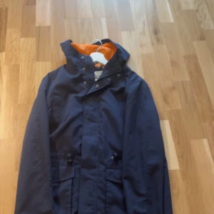 Mörkblå vindjacka med orange foder - Snygg mörkblå vindjacka med huva och orange foder. Jackan har dragsko i midjan, knappar och dragkedja framtill samt praktiska fickor. Perfekt för blåsiga dagar och enkel att matcha med olika outfits.