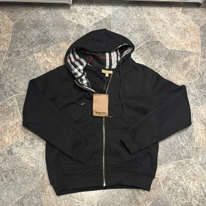 Svart Burberry hoodie med dragkedja - Svart hoodie från Burberry i bomull med klassiskt rutigt foder i huvan och broderad logga på bröstet. Tröjan har hel dragkedja framtill, snörning i huvan och ribbade muddar. Perfekt för en stilren och avslappnad look.