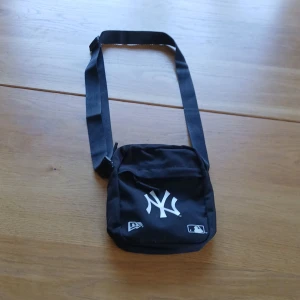Svart New Yorkyankees axelväska - Säljer en svart axelväska från New Era med New York Yankees-logga framtill. Väskan har justerbar rem, dragkedja och är tillverkad i slitstark polyester. Perfekt storlek för mobilen, plånboken och andra småprylar. MLB-logga på både fram- och baksidan. Sparsamt använd. skriv om du har några frågor. Pris kan diskuteras.