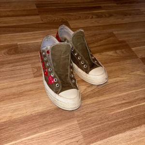 Converse Play x CDG sneakers olivgrön - Säljer ett par Converse Play x Comme des Garçons sneakers i olivgrönt canvasmaterial med vita sulor och röda hjärtan på sidorna. Klassisk rund tå och platt sula, utan snörning. Perfekta för dig som gillar streetstyle och unika detaljer.