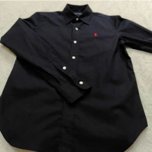 Svart skjorta från Ralph Lauren - Klassisk svart skjorta från Ralph Lauren med lång ärm och vit knappstängning. Skjortan har den ikoniska röda broderade loggan på bröstet och är tillverkad i mjuk bomull. Perfekt för dig som gillar stilrena och tidlösa plagg.