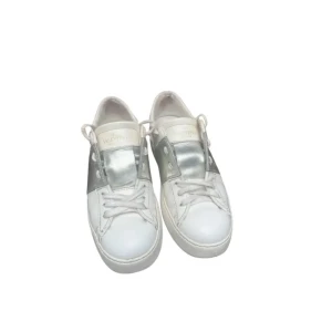 Valentino opens i vitt och silver - Snygga sneakers från Valentino Garavani i vitt skinn med bred silverfärgad detalj över snörningen. Klassisk rund tå, vita snören och ikoniska nitar baktill på sulan. Perfekta för dig som vill sticka ut med en lyxig touch. Kvitto, box och tags medföljer