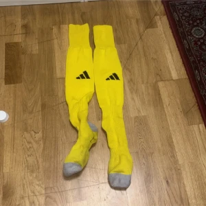 Gula Adidas fotbollsstrumpor - Långa gula fotbollsstrumpor från Adidas med svart logga på framsidan och grå förstärkning vid tår och häl. Perfekta för match eller träning, klassisk sportig look och bekväm passform.