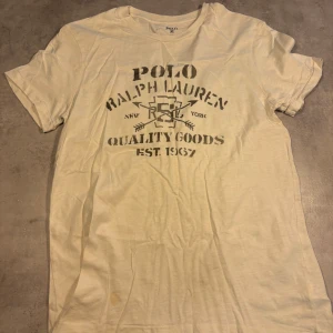 Vit t-shirt Polo Ralph Lauren M - Vit t-shirt från Polo Ralph Lauren i storlek M. Tryck på bröstet med text och grafiska detaljer, klassisk rund hals och korta ärmar. Skön och mjuk bomull, perfekt för chill dagar. Snygg vintage-vibe med coolt tryck och relaxed passform.
