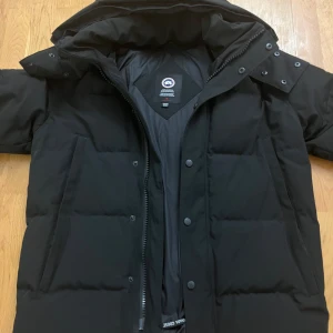 Svart jacka från Canada Goose black label  - Säljer en svart dunjacka från Canada Goose med huva och stora fickor framtill. Jackan har en rejäl dragkedja, tryckknappar och klassisk patch på ärmen. Perfekt för kalla dagar och riktigt snygg streetstil.