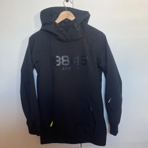 Svart jacka från 8848 Altitude - Svart jacka från 8848 Altitude med stor logga framtill. Tröjan har huva med snörning, långa ärmar och praktisk ficka framtill. Perfekt för en skidtur, har en sportig vibe. Materialet känns slitstarkt och är troligtvis polyester. Knappt använd.