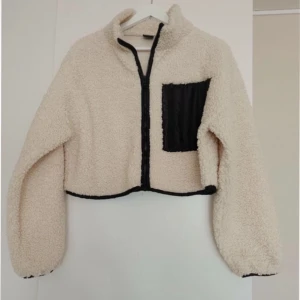 Beige teddyjacka med svart detalj - Säljer en beige teddyjacka med croppad passform och svart dragkedja. Jackan har en svart bröstficka i kontrastmaterial och svarta detaljer längs kanterna. Perfekt för dig som vill ha en mysig och trendig look. Större i storleken ett hål vid fickan