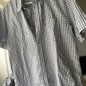 Blårutig Peak Performance skjorta - Snygg kortärmad skjorta från Peak Performance i blått och vitt rutmönster. Skjortan har klassisk krage, bröstficka och knäppning framtill. Perfekt för en avslappnad och stilren look under varmare dagar.