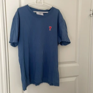 Blå t-shirt från Ami med hjärtdetalj - Snygg blå t-shirt från Ami Alexandre Mattiussi med klassisk rund hals och korta ärmar. På bröstet sitter Ami:s ikoniska broderade hjärta med ett A i rött. T-shirten är gjord i mjuk bomull och har en normal passform, perfekt för en chill och stilren look.