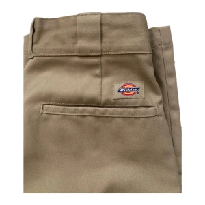 Beige Dickies byxor - Klassiska Dickies 874 chinos i beige med rak passform och normal midja. Byxorna har diskret Dickies-logga ovanför bakfickan, bälteshällor och dragkedja. Tillverkade i slitstarkt bomullsmaterial med en clean och tidlös look. Har en grå rand på ena innerlåret men syns inte så väl.